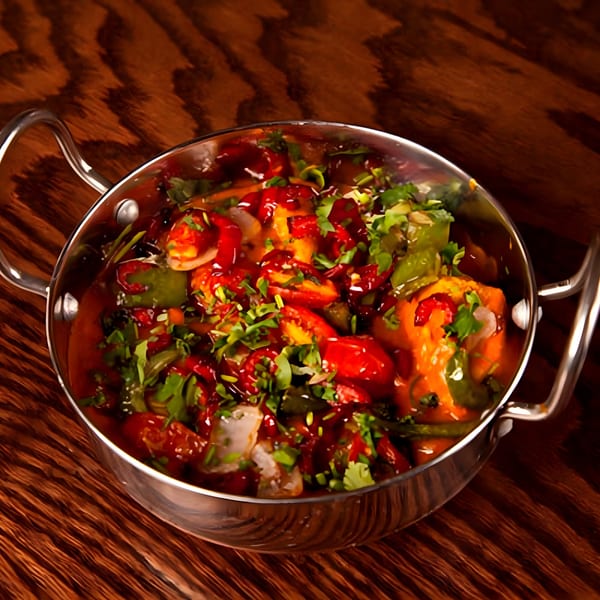 Indian dish with red sauce, green peppers, and cilantro in a metal serving bowl on a wooden table.