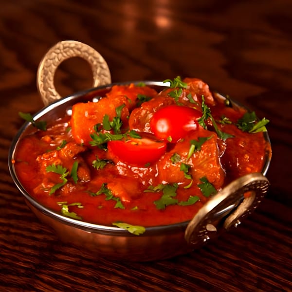 A copper karahi bowl filled with vibrant red Indian curry, garnished with fresh cilantro and cherry tomatoes.