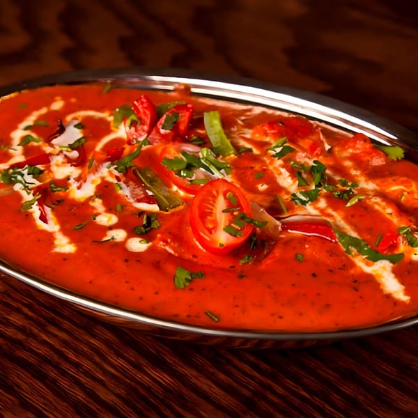 A vibrant Indian curry in a silver dish, garnished with fresh herbs, tomatoes, and cream swirls.