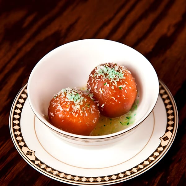 Two gulab jamun, Indian fried dough balls, in a white bowl with decorative saucer on a wooden table.